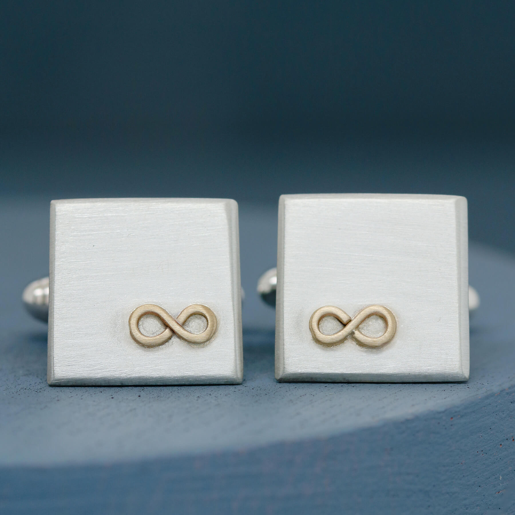 Pair of gold infinity Cufflinks personalised, face on to show the raised 9ct gold detail against matte silver, photographed on a deep blue background.