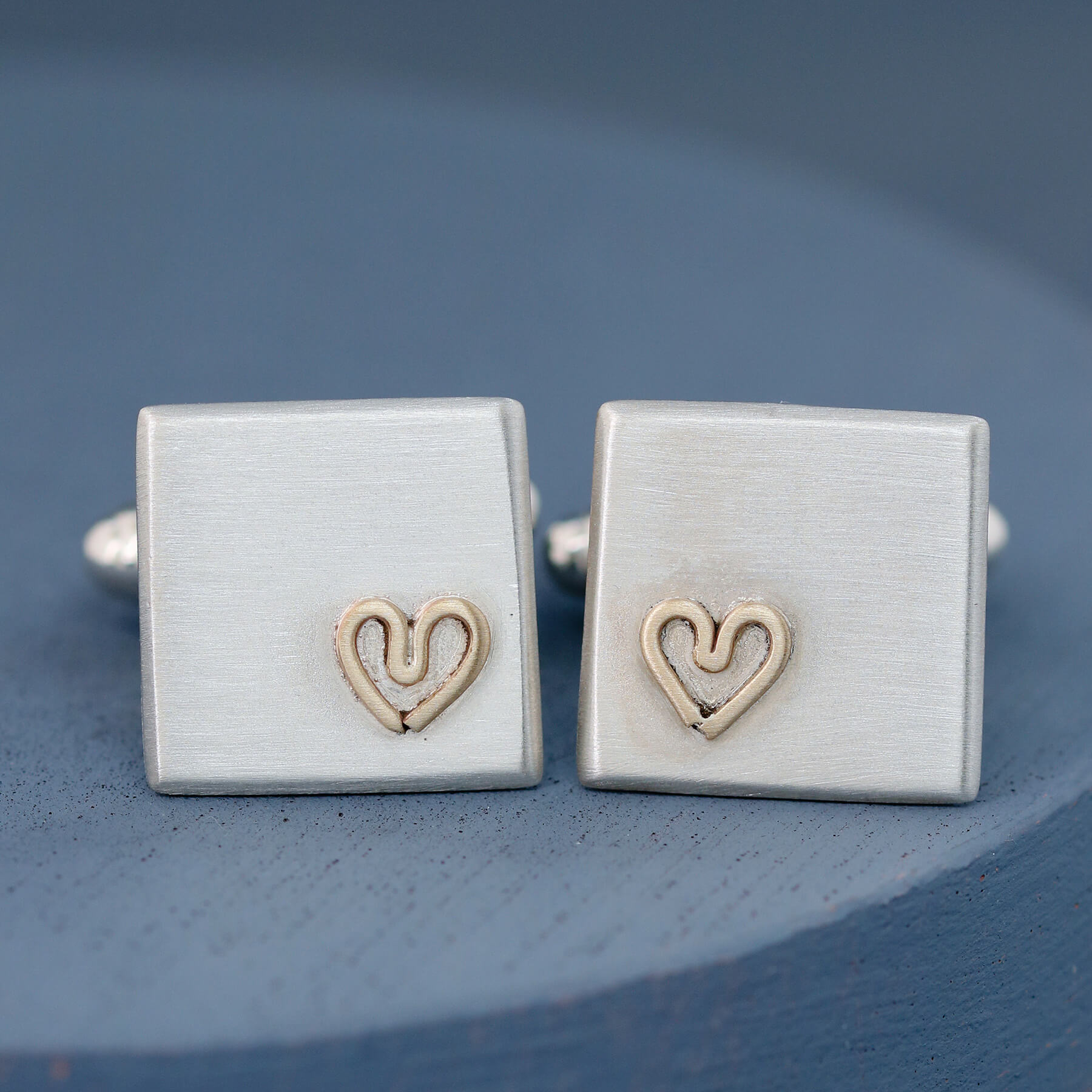 Pair of sterling silver cufflinks with a small 9ct gold heart on each, photographed on a deep blue background — Custom cufflinks for groom.