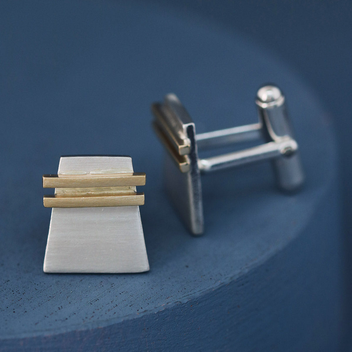 Detailed close-up of silver and gold cufflink face on a deep blue background, emphasising craftsmanship; refined luxury cufflinks for Father’s Day or anniversaries.