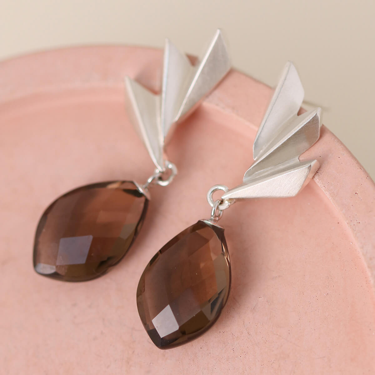 Smoky quartz earrings laid flat on a pink dish with dramatic gemstone facets and geometric silver detailing; luxurious art deco statement earrings for her.