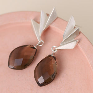Smoky quartz earrings laid flat on a pink dish with dramatic gemstone facets and geometric silver detailing; luxurious art deco statement earrings for her.