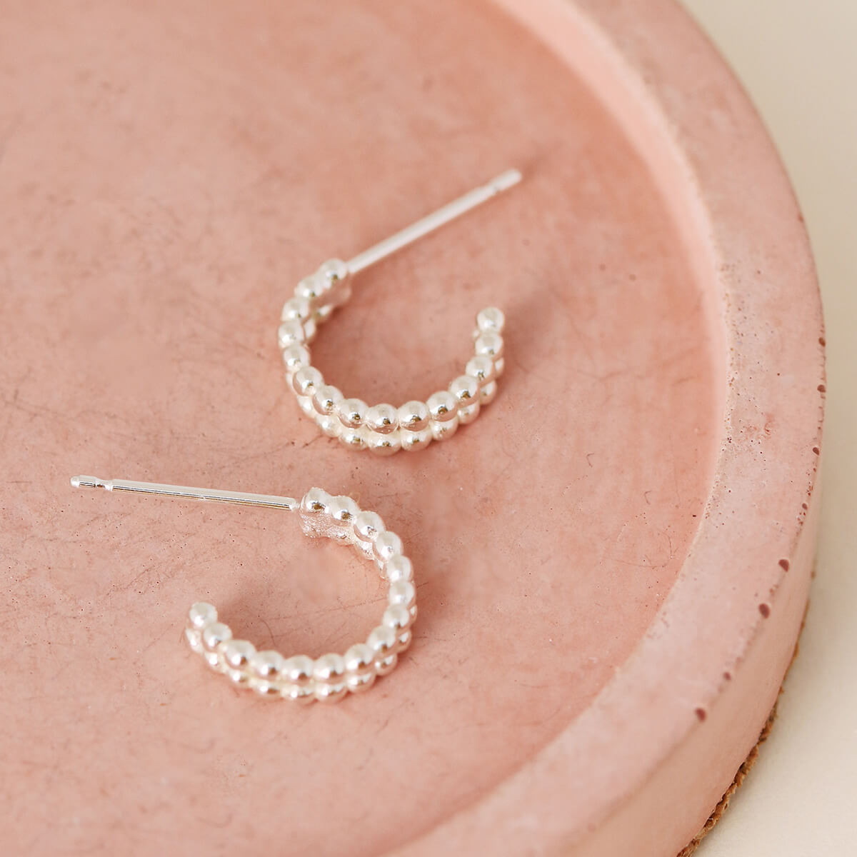 Close up of small huggie hoop earrings in sterling silver on a pink dish, everyday minimalist jewellery