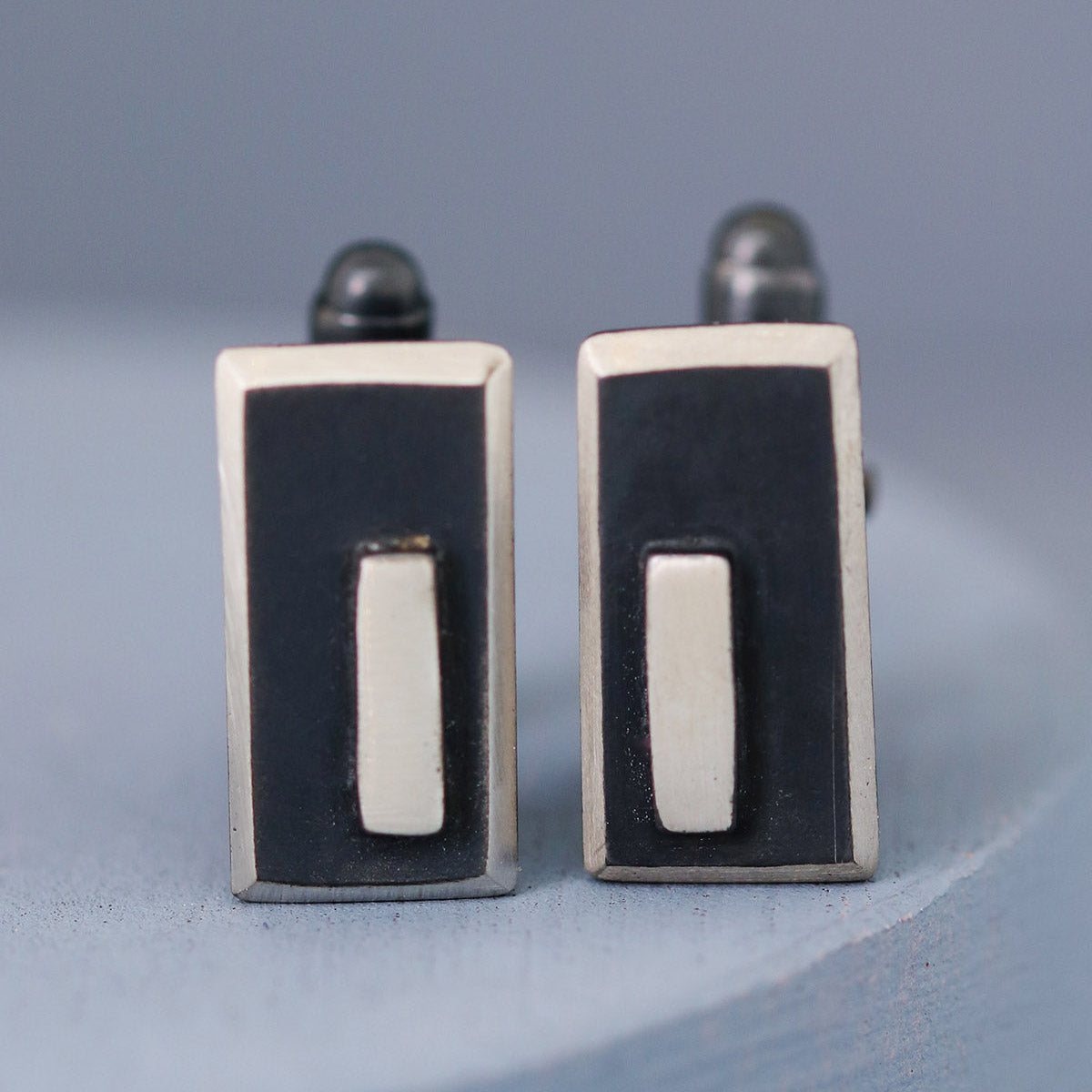 Close-up of one cufflink in focus with the other softly blurred behind, displaying the bold shape and texture of these black and silver cufflinks.