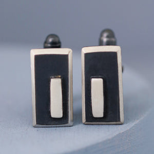Close-up of one cufflink in focus with the other softly blurred behind, displaying the bold shape and texture of these black and silver cufflinks.