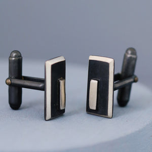 Handmade black and silver cufflinks photographed flat on a deep blue surface, showcasing their clean geometric lines and contemporary design.