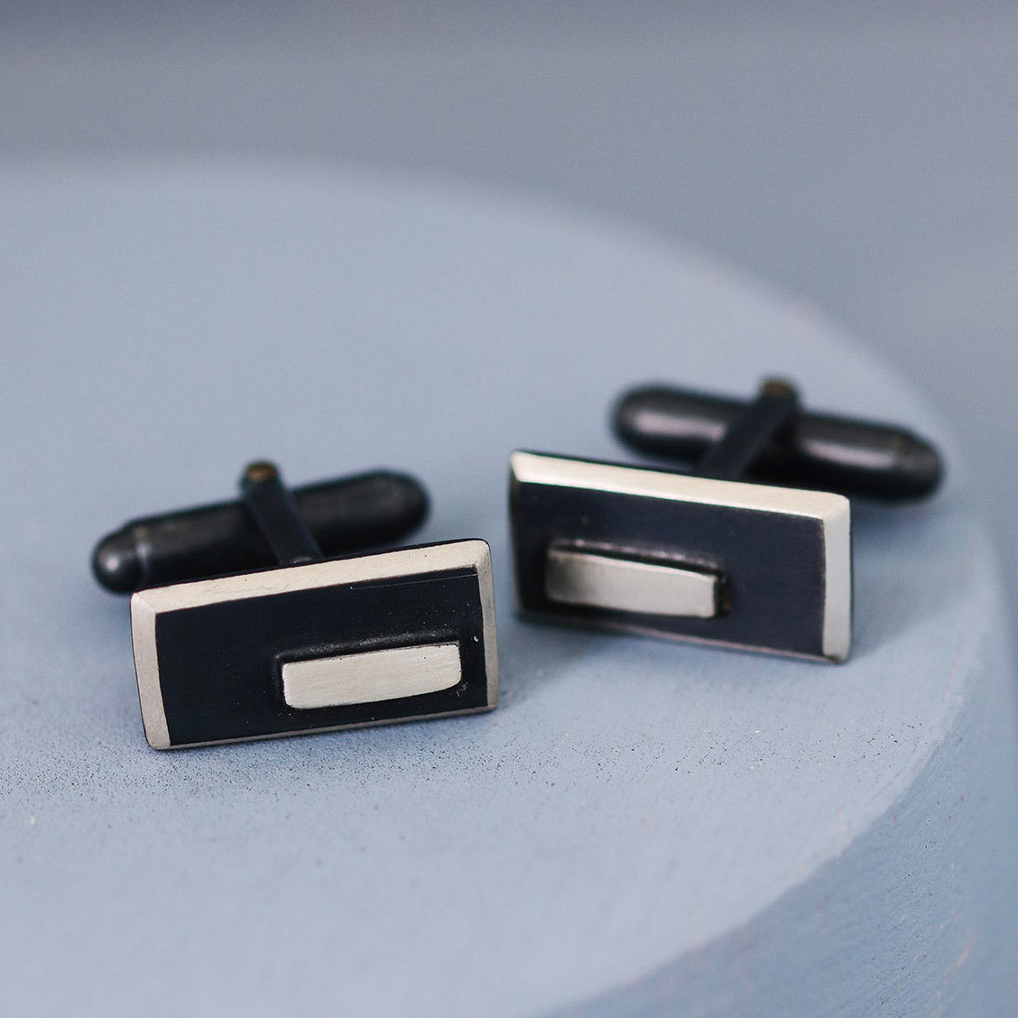 Pair of black and silver cufflinks side by side on a deep blue background, highlighting the striking contrast between the oxidised black and polished silver finish.