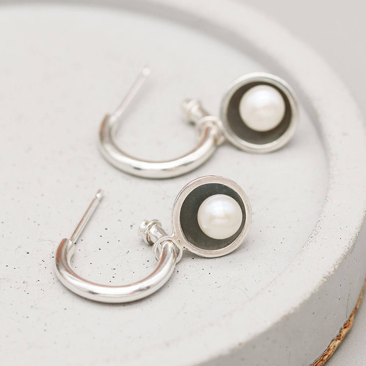 Black pearl hoop earrings handcrafted in sterling silver with removable freshwater pearl charms displayed on a grey ceramic dish.