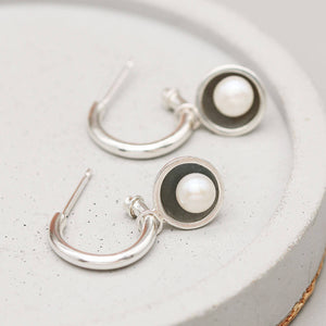 Black pearl hoop earrings handcrafted in sterling silver with removable freshwater pearl charms displayed on a grey ceramic dish.