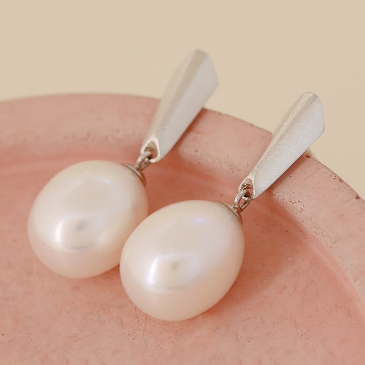 Silver dangle pearl earrings arranged on a pink dish to showcase the hand-carved geometric silver accents and natural pearl sheen.