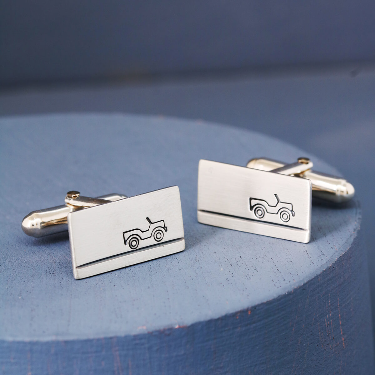 Silver land rover cufflinks with matte brushed finish displayed on blue background, thoughtful anniversary gift for husband who loves cars.