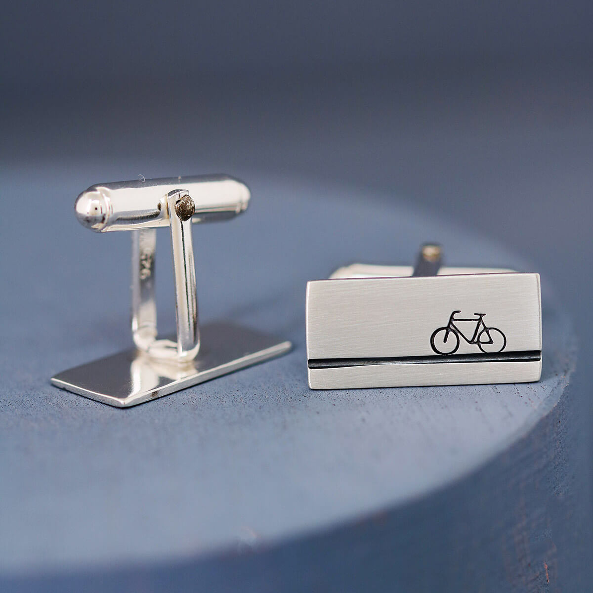 Side view of matte brushed finish cycling cufflinks on a deep blue background, modern sterling silver gift for bicycle lovers and bike enthusiasts.