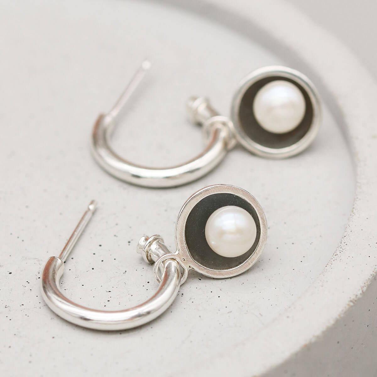 Handmade black pearl hoop earrings with removable freshwater pearl charms displayed on a grey ceramic dish.