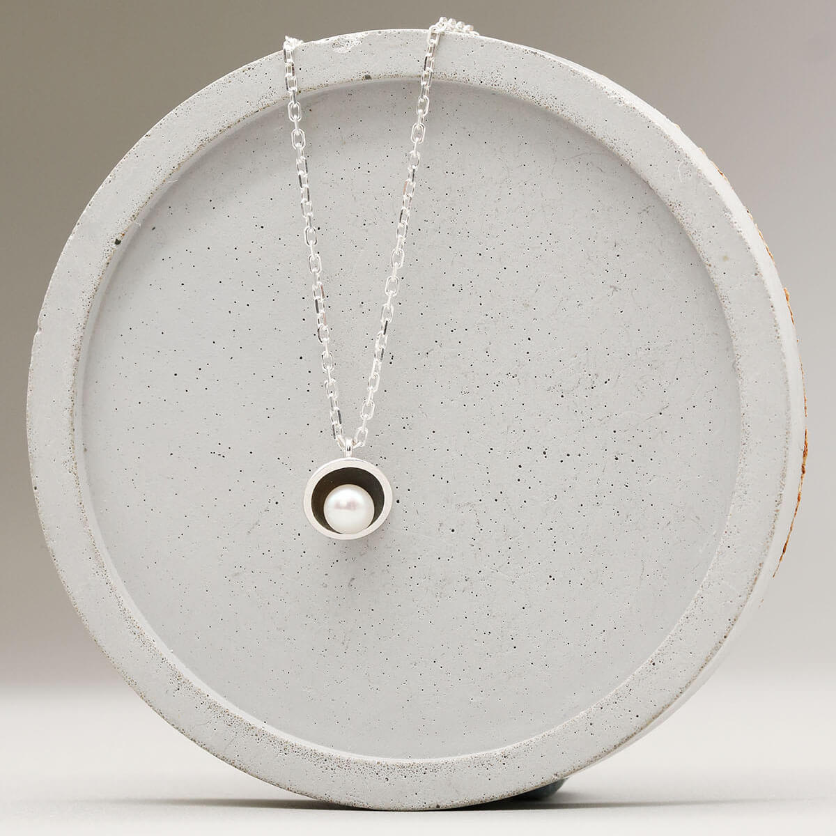 Contemporary black pearl pendant necklace with a handcrafted sterling silver setting and freshwater pearl displayed on a grey dish.