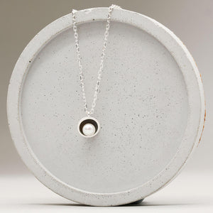 Contemporary black pearl pendant necklace with a handcrafted sterling silver setting and freshwater pearl displayed on a grey dish.