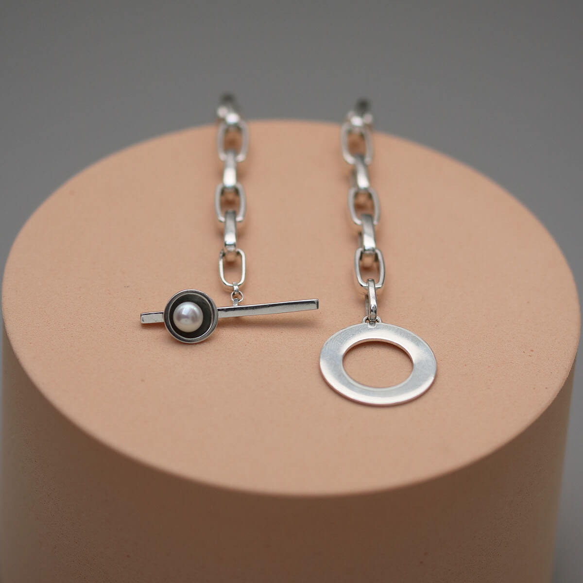 Sterling silver pearl statement necklace displayed on a pink circular plinth, showcasing the geometric T-bar design and chunky chain.