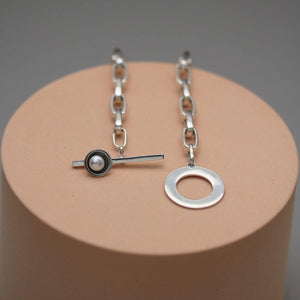 Sterling silver pearl statement necklace displayed on a pink circular plinth, showcasing the geometric T-bar design and chunky chain.