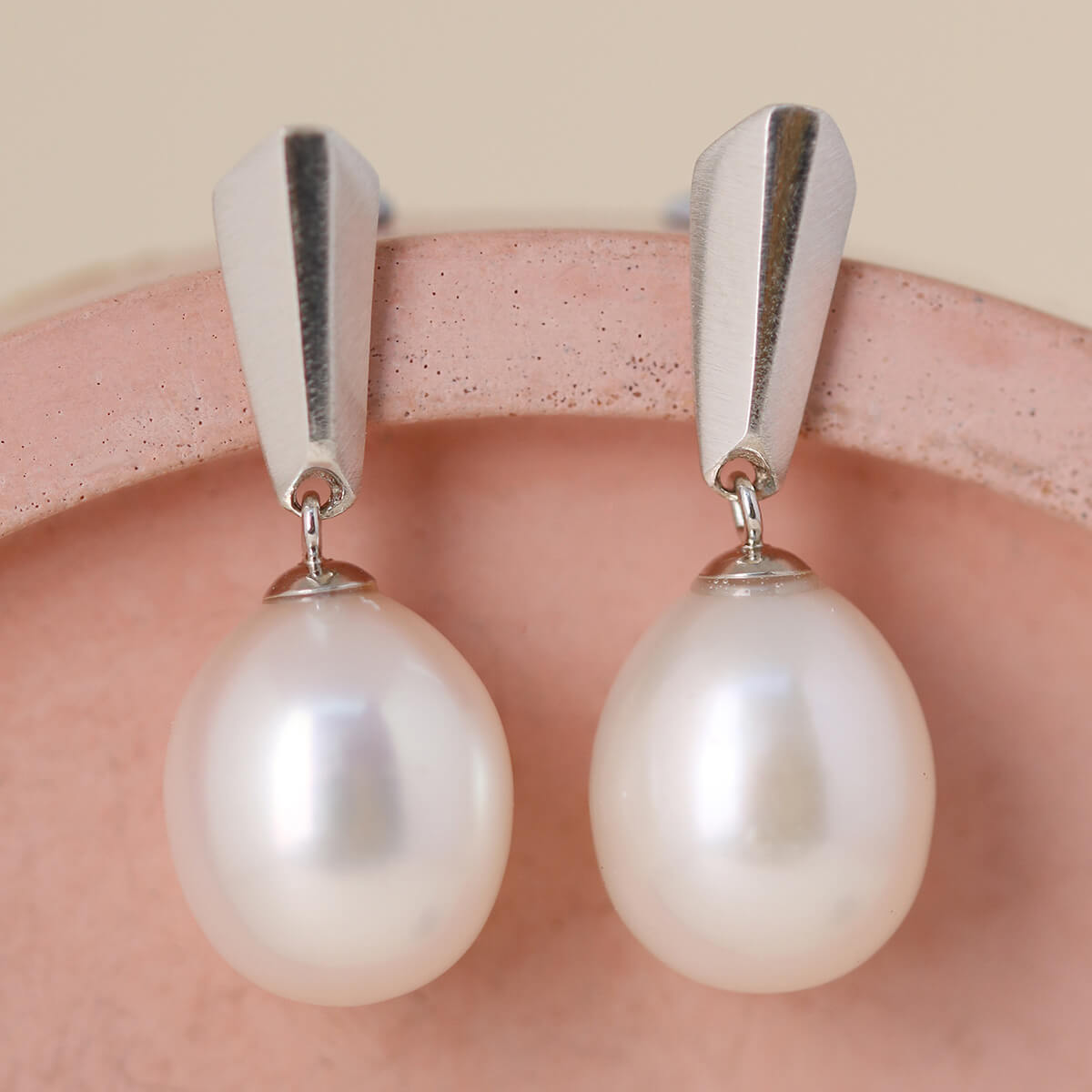 Side angle of sterling silver dangle pearl earrings on a pink dish, revealing the gentle movement and craftsmanship.