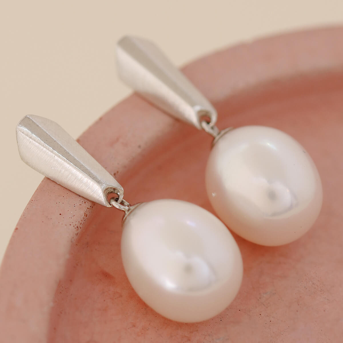 Sterling silver dangle pearl earrings displayed on a pink dish, showing the geometric silver detail and large lustrous pearls.