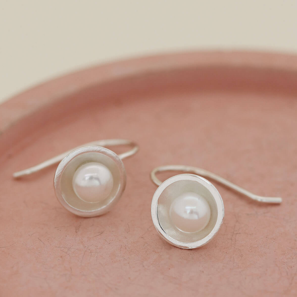 Contemporary sterling silver pearl drop earrings handcrafted with genuine freshwater pearls on a pink ceramic dish.
