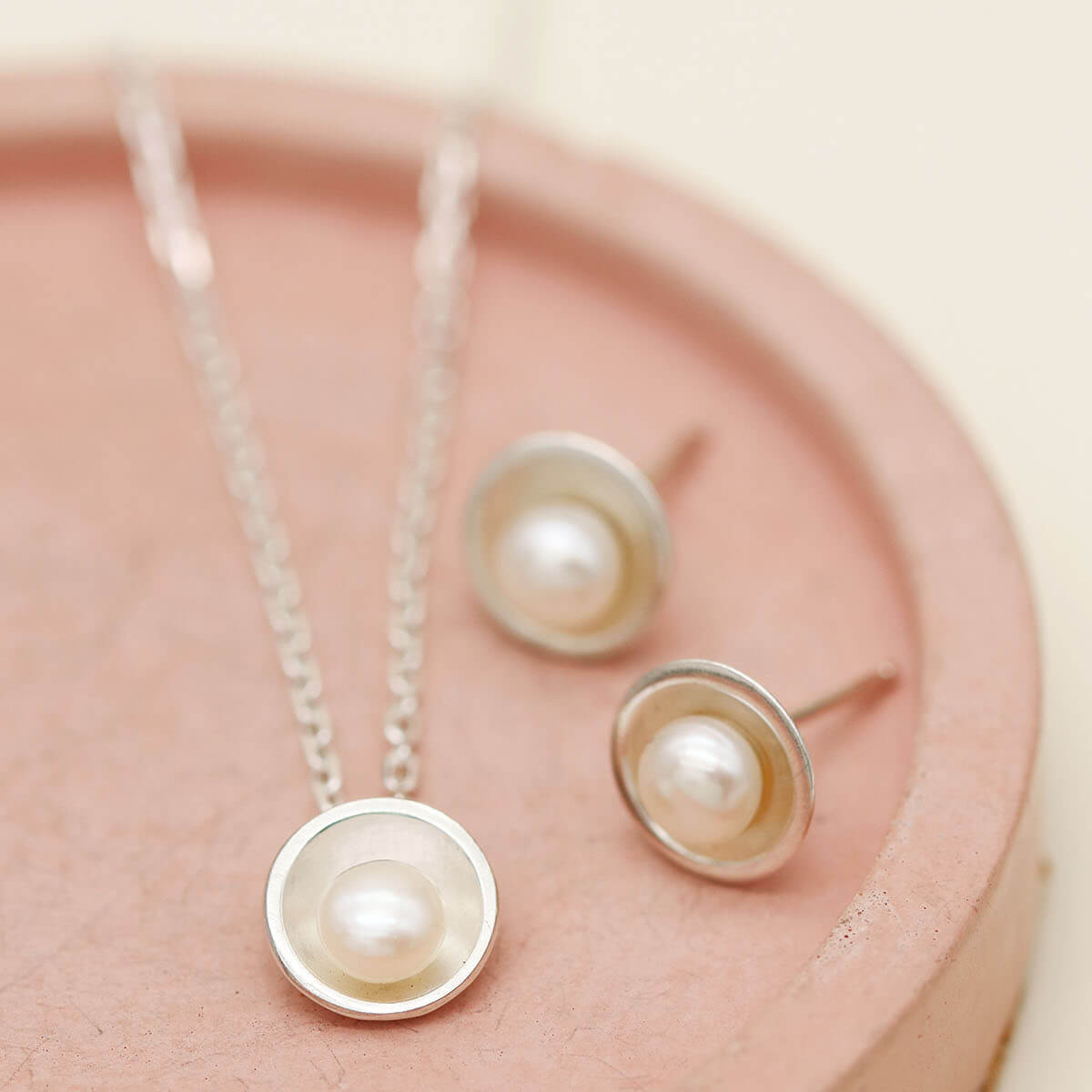 Handmade silver pearl pendant necklace shown alongside matching sterling silver pearl stud earrings on a pink dish.