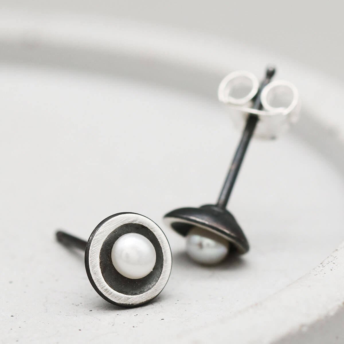 Tiny pearl stud earrings handcrafted from oxidised sterling silver with genuine freshwater pearls on a dark grey dish.