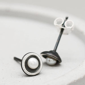 Tiny pearl stud earrings handcrafted from oxidised sterling silver with genuine freshwater pearls on a dark grey dish.