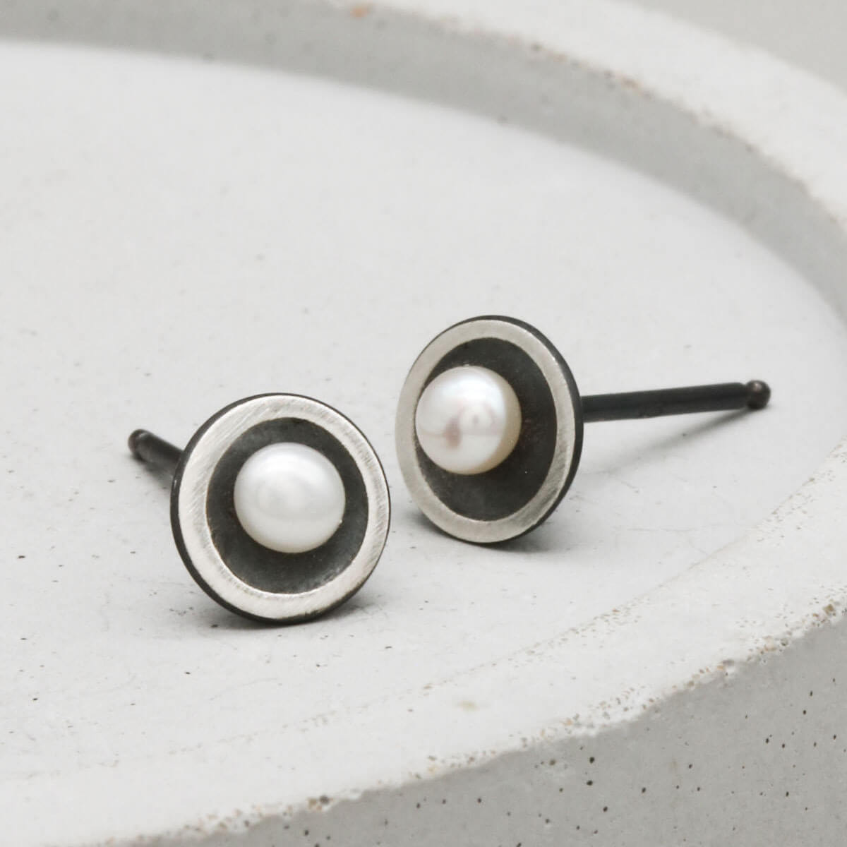 Handmade Petite Tiny Pearl Stud Earrings in oxidised sterling silver with genuine freshwater pearls displayed on a dark grey ceramic dish.