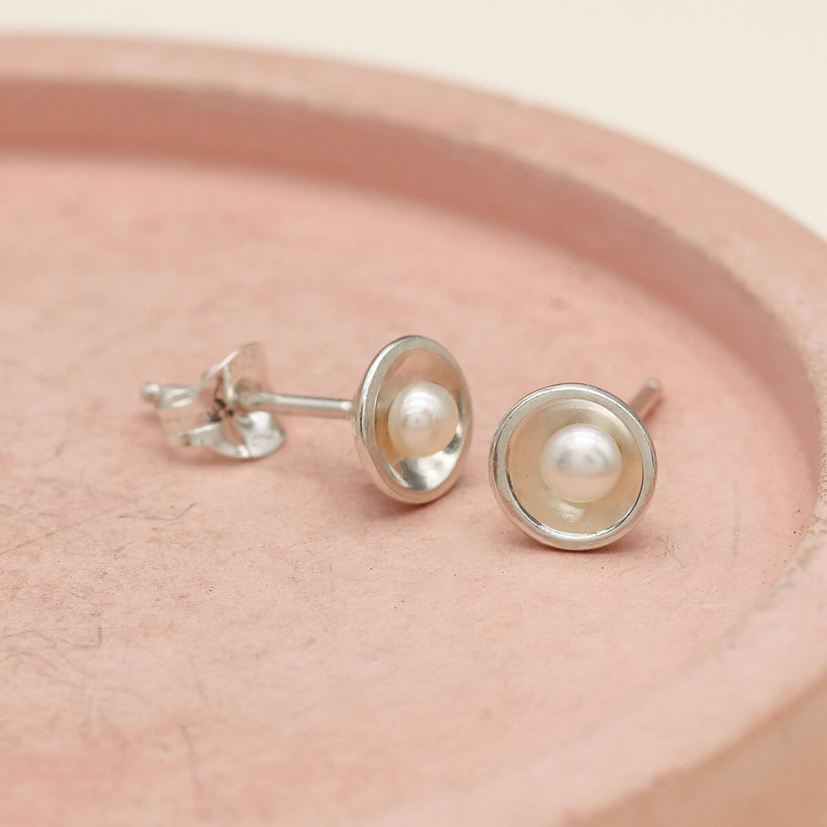 Delicate sterling silver pearl stud earrings on a pink ceramic dish