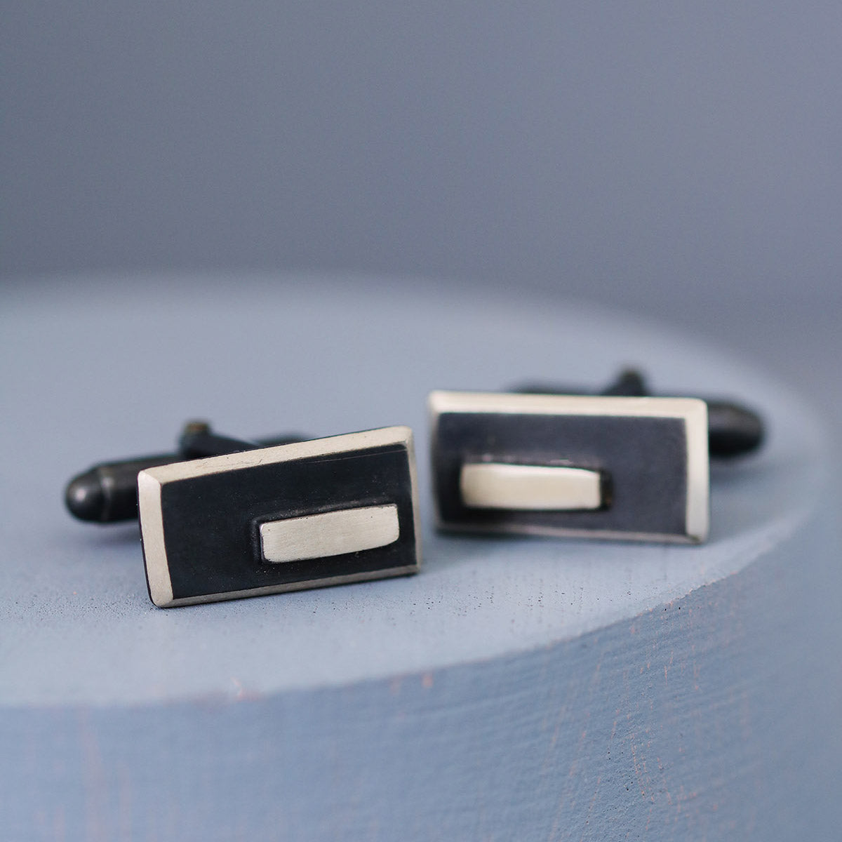 Angled view of black and silver cufflinks catching the light on a deep blue background, showing their stylish contrast and modern elegance.