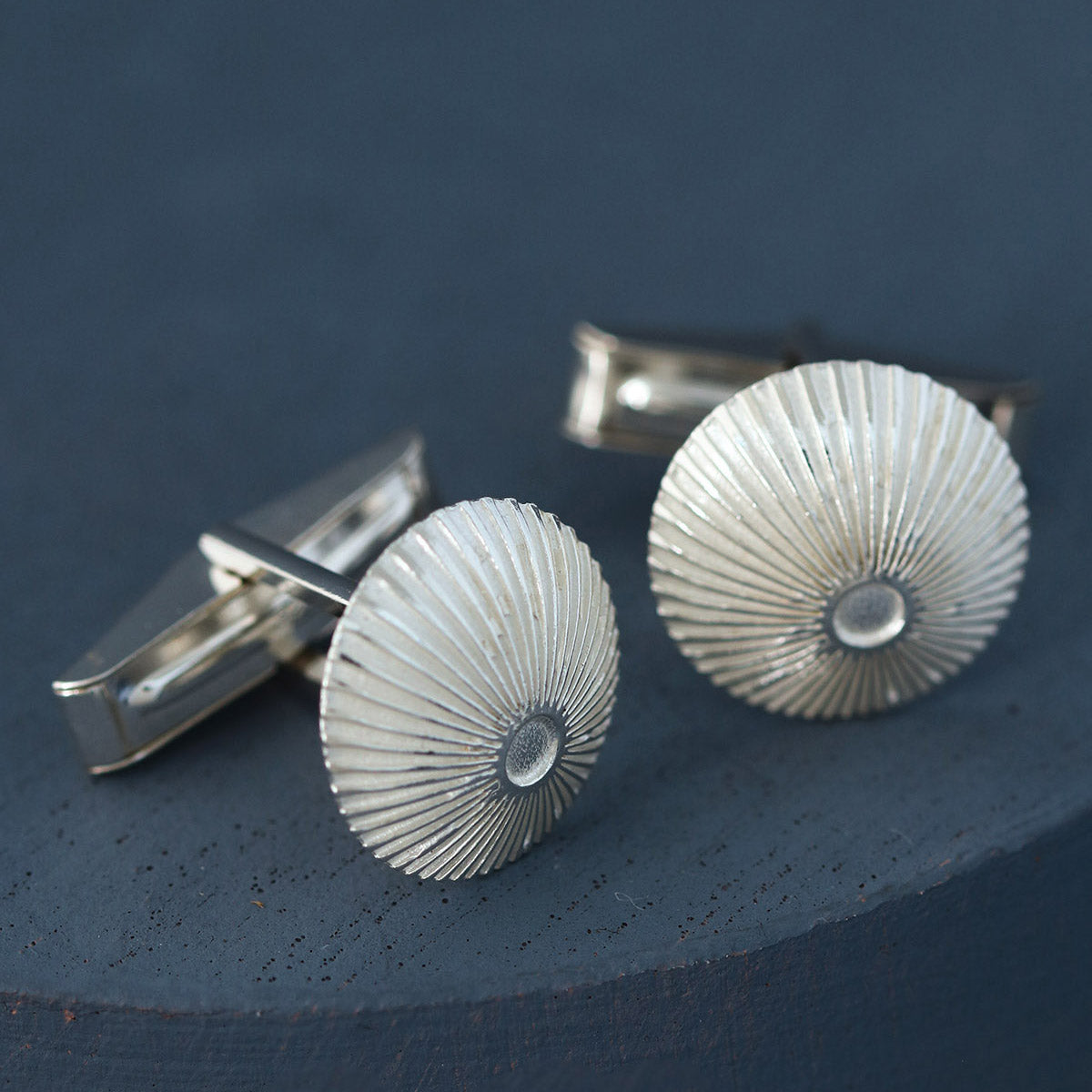 Round cufflinks in sterling silver with crisp engraved pattern, styled on deep blue background – unique artisan gift for cyclist or commuter.