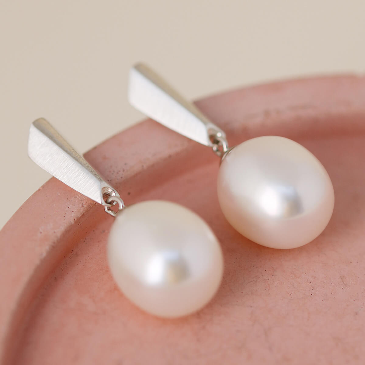 Close-up of silver dangle pearl earrings on a soft pink dish highlighting the Art Deco inspired silver top and smooth 12mm pearls.