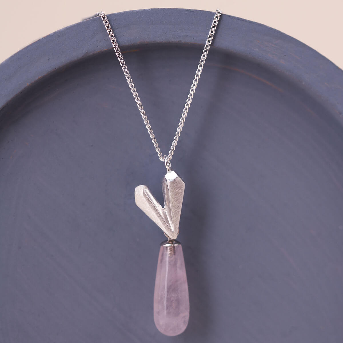 Sterling silver heart necklace with pink quartz heart pendant laid on a blue dish, a delicate handmade heart jewellery design.