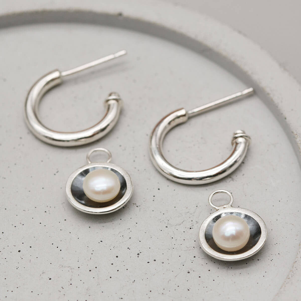 Handmade hoop earrings with pearl drop shown with removable freshwater pearl charms on a grey dish.