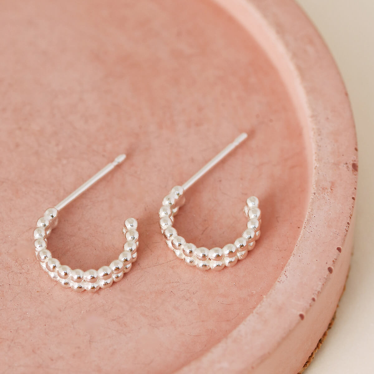 Minimalist sterling silver huggie earrings laid on a pink dish, small huggie hoop earrings