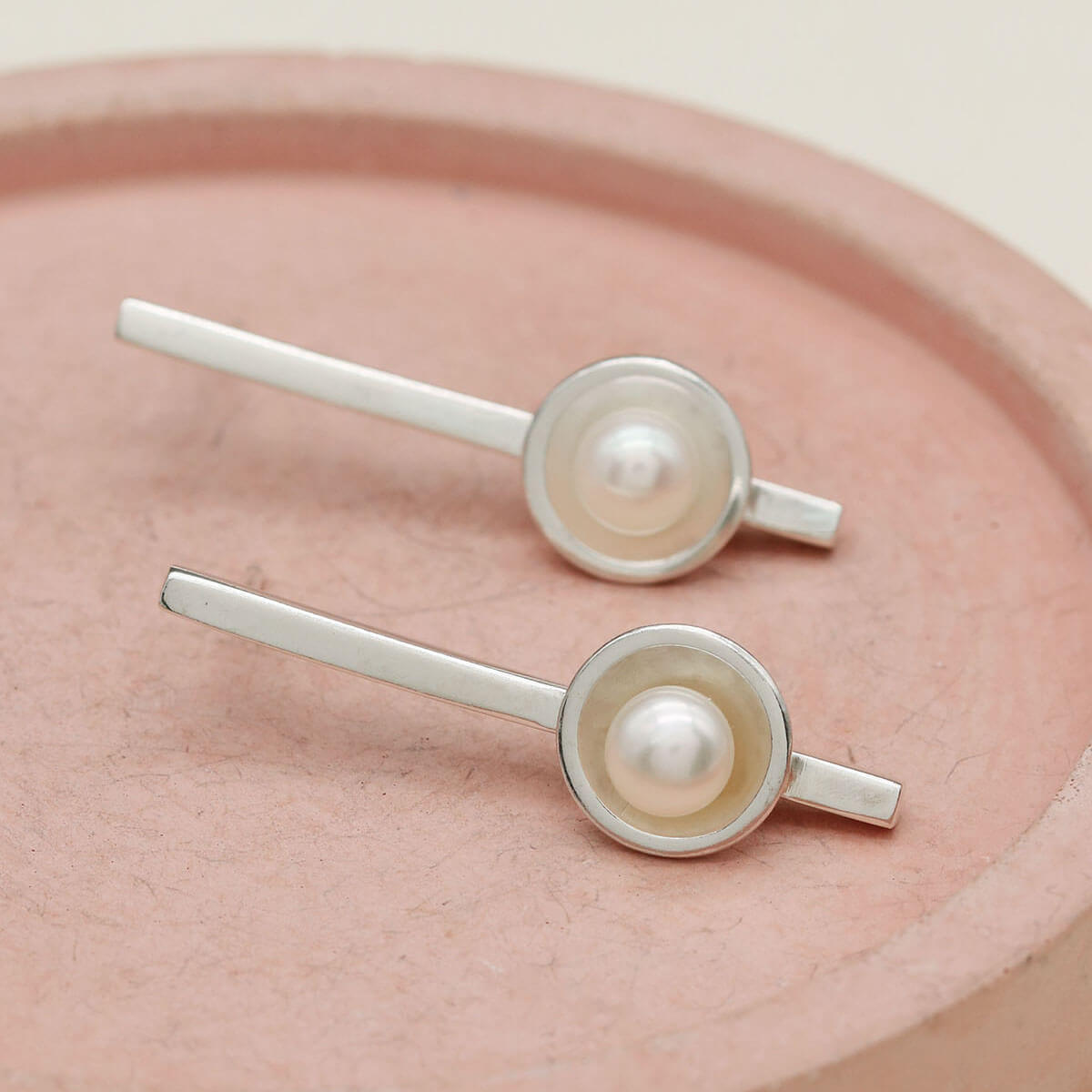 Contemporary sterling silver long pearl earrings with handcrafted freshwater pearl settings displayed on a pink dish.