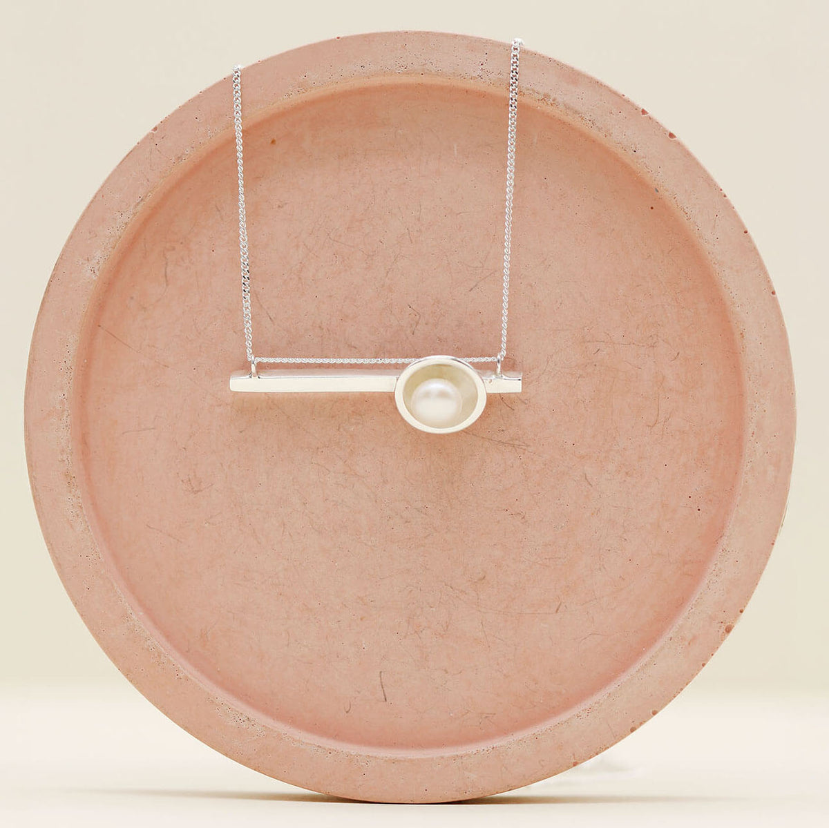 Handmade sterling silver geometric pearl necklace with a horizontal bar pendant and genuine freshwater pearl on a pink ceramic dish.