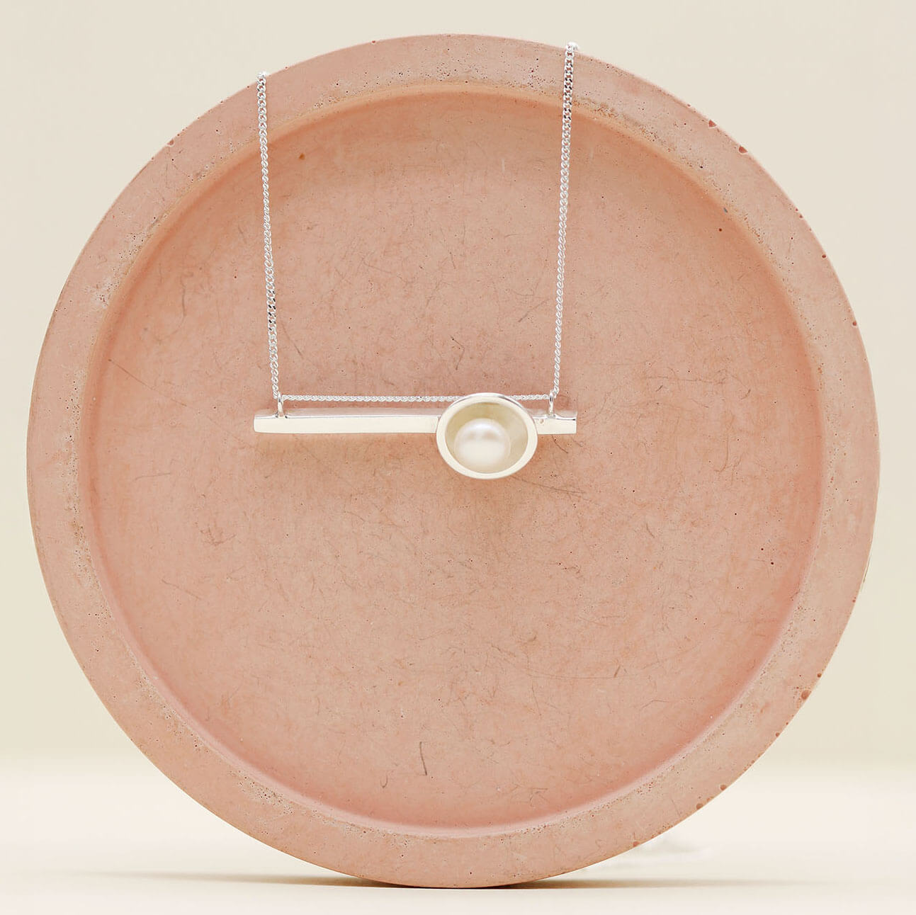 Handmade sterling silver geometric pearl necklace with a horizontal bar pendant and genuine freshwater pearl on a pink ceramic dish.