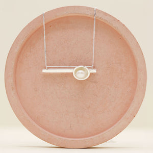 Handmade sterling silver geometric pearl necklace with a horizontal bar pendant and genuine freshwater pearl on a pink ceramic dish.