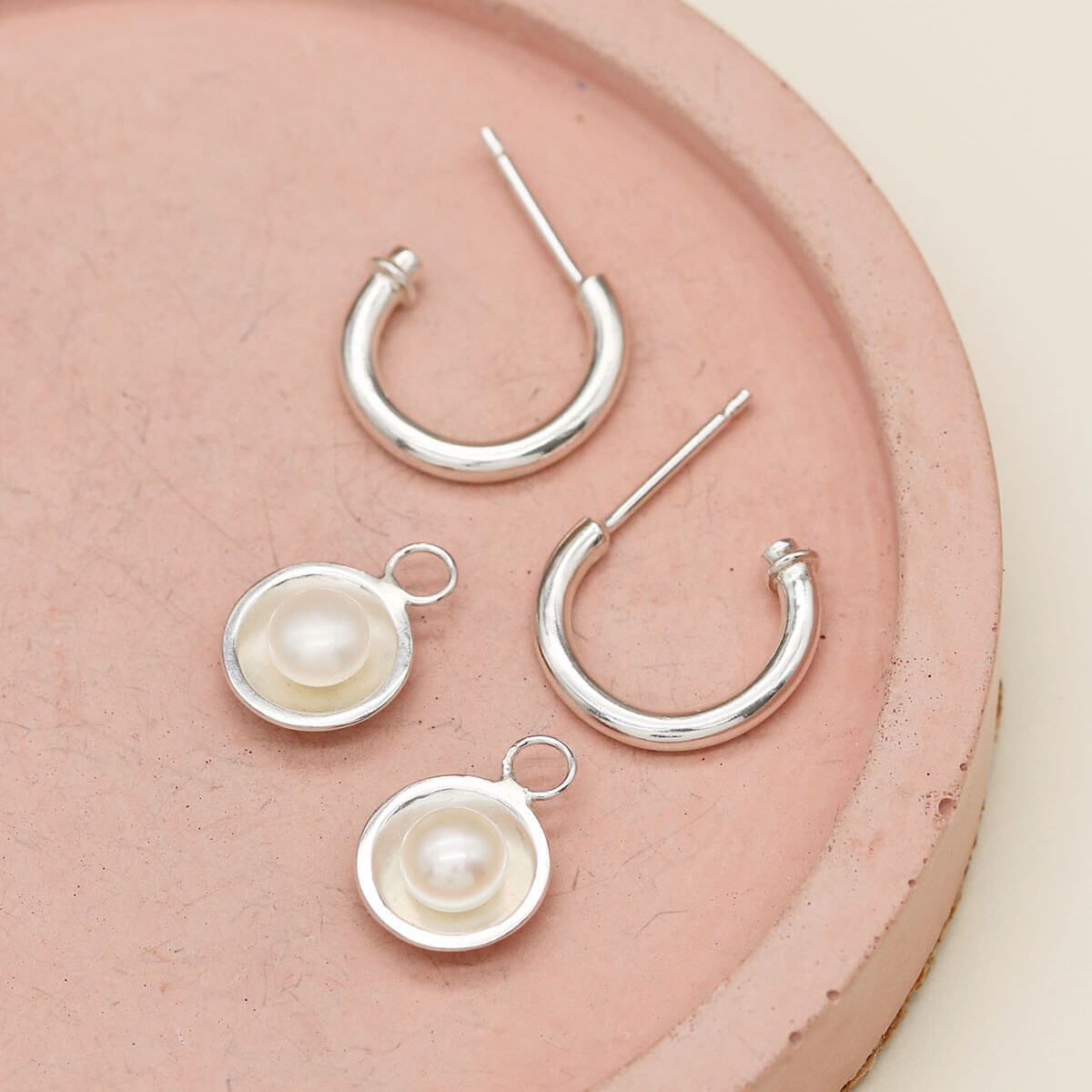 Sterling silver hoop earrings with pearls shown with detachable freshwater pearl charms on a pink ceramic dish.